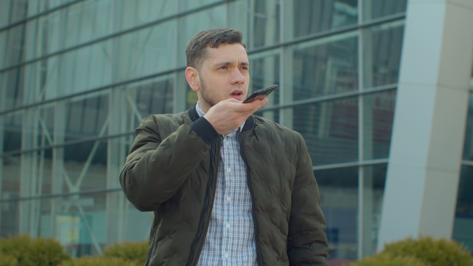 junger Mann draussen vor Glasgebäude, hält Handy an den Mund, Mund offen. Trägt Jacke und hellblau-kariertes Hemd.