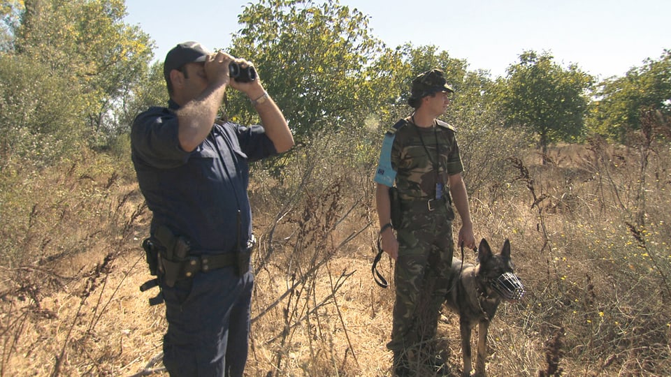 Zwei Grenzpolizisten mit Feldstecher und Hund in trockener Landschaft.