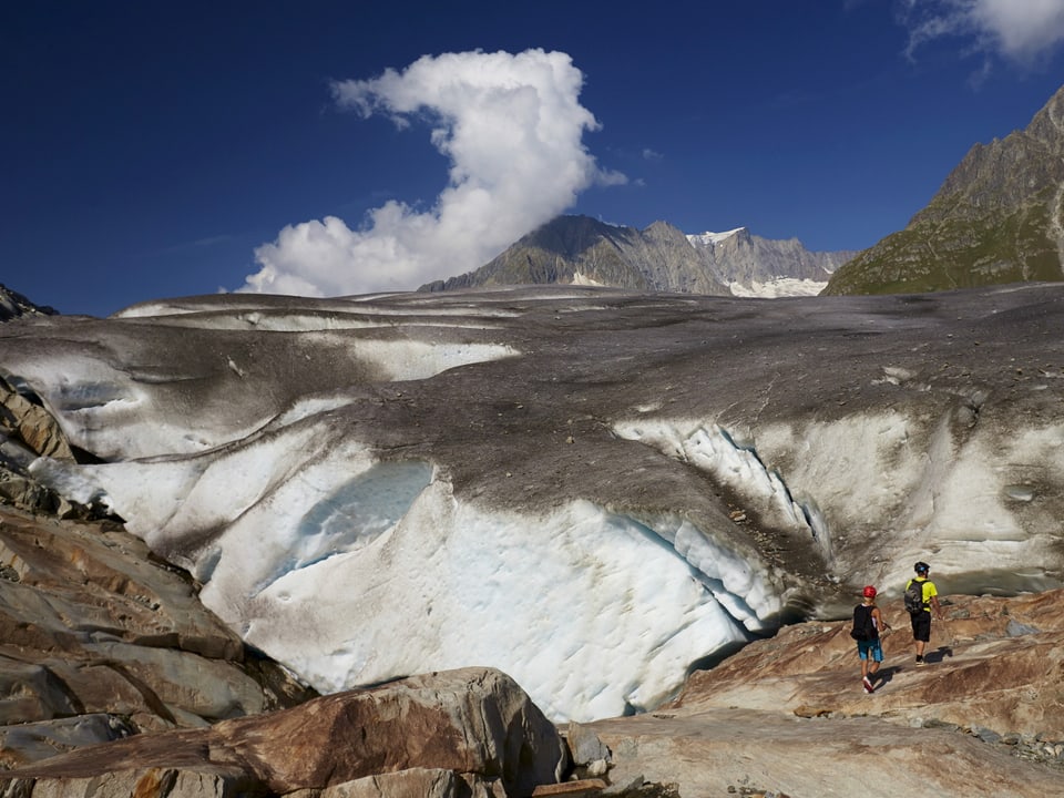 Gletscherzunge von unten, zwei Wanderer auf den Felsen daneben unterwegs.