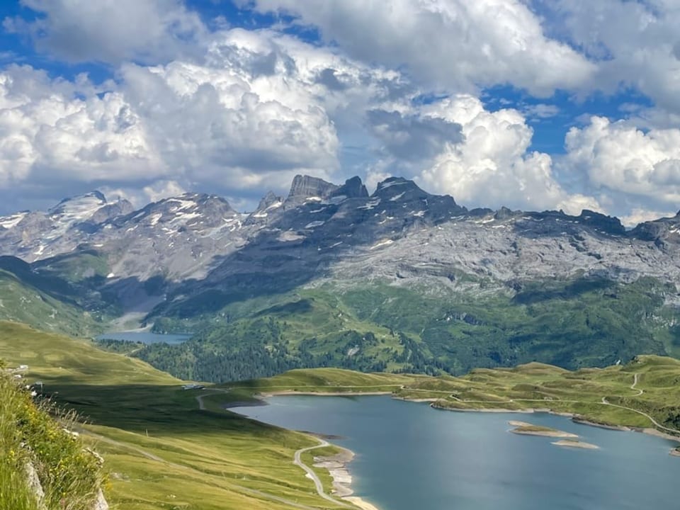 Bergsee mit grüner Alpwiese, im Hintergrund felsige Berge mit Quellwolken am Himmel.