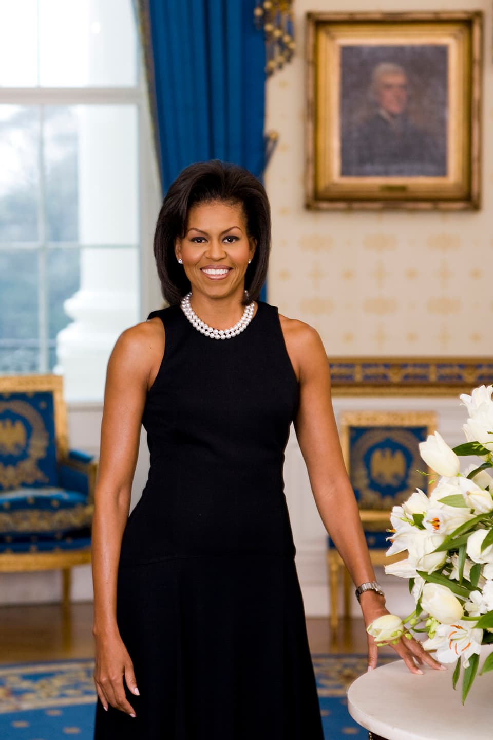 Michelle Obama an einem Tisch, der mit Blumen geschmückt ist, stehend in schwarzem Kleid und Perlenkette.
