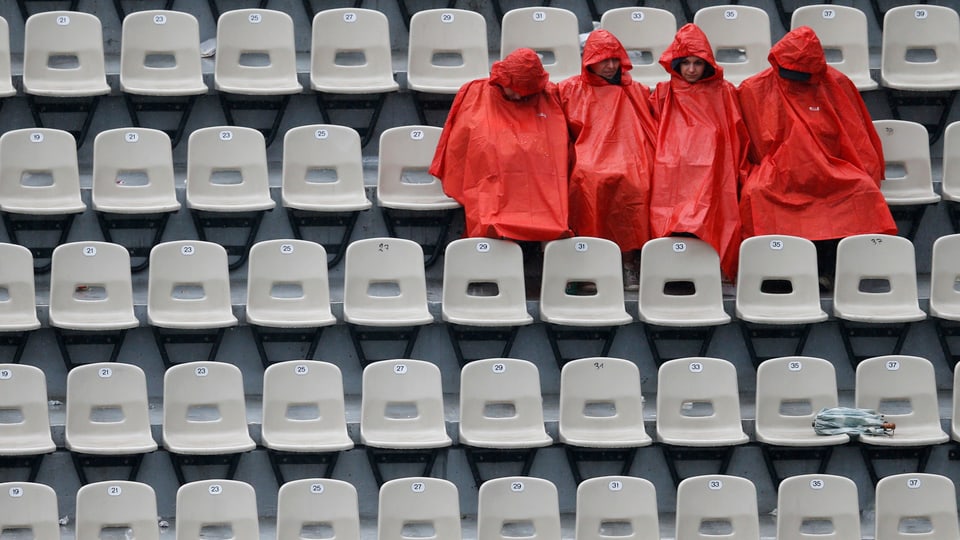 Vier Menschen sitzen in roten Regenjacken auf Zuschauertribünen.