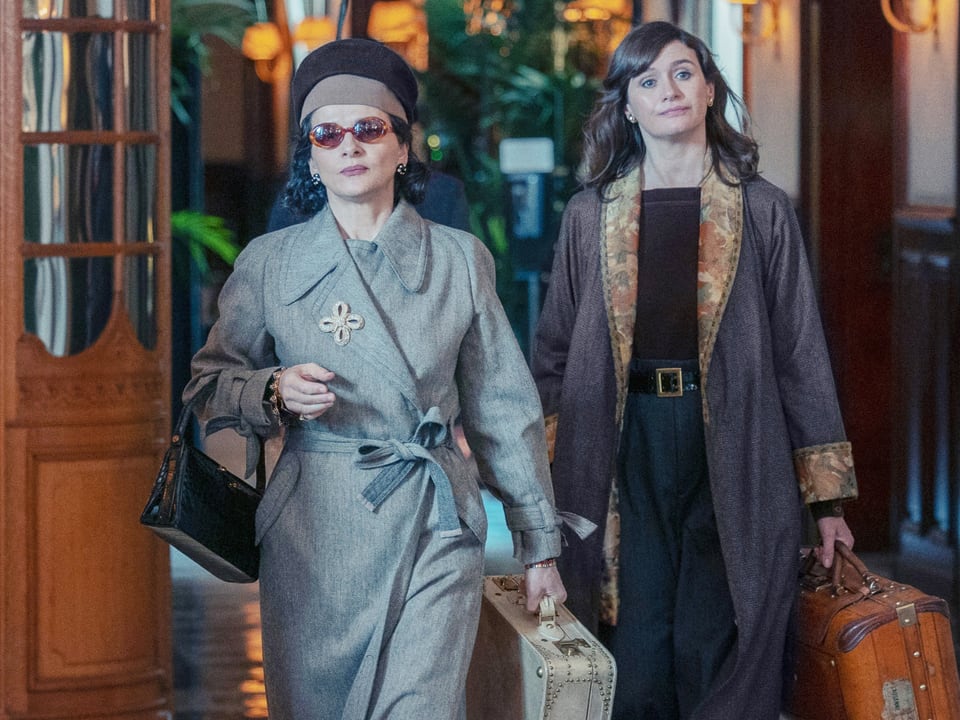 Zwei Frauen, angezogen mit Mantel, die Frau links mit Hut und Sonnenbrille. Beide tragen Tasche und Koffer.