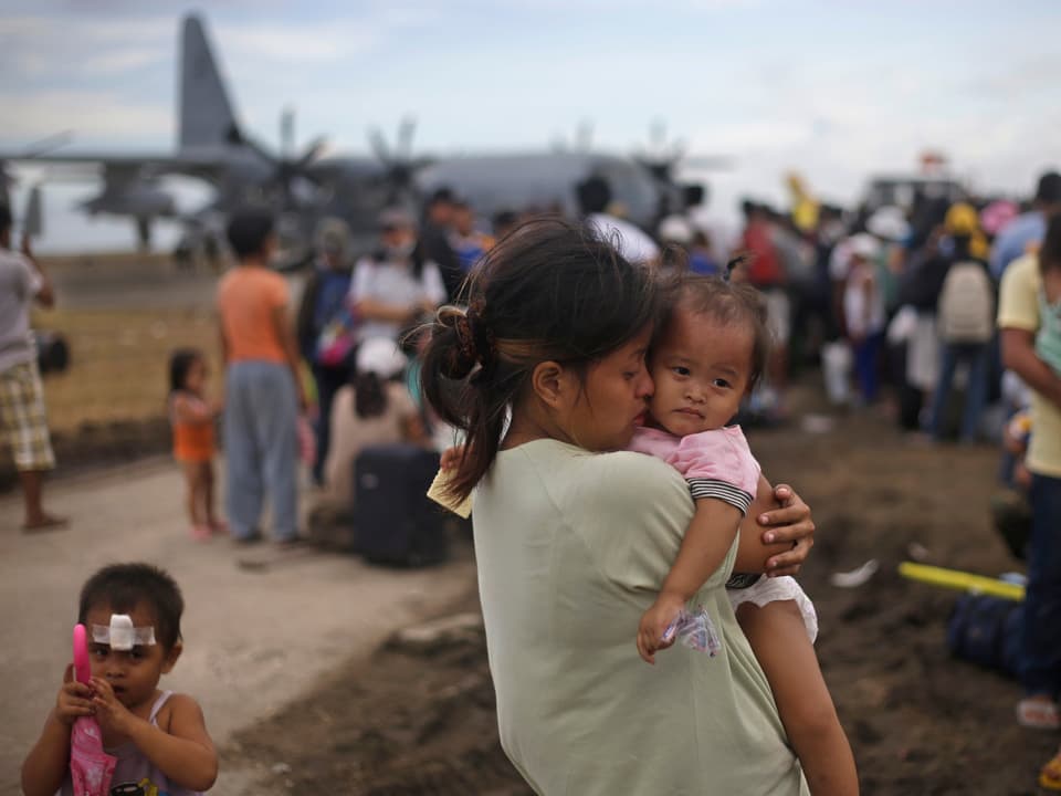 Eine Frau hält ihr Kind im Arm und steht vor einer Gruppe wartender Menschen vor einem Hilfs-Flugzeug.