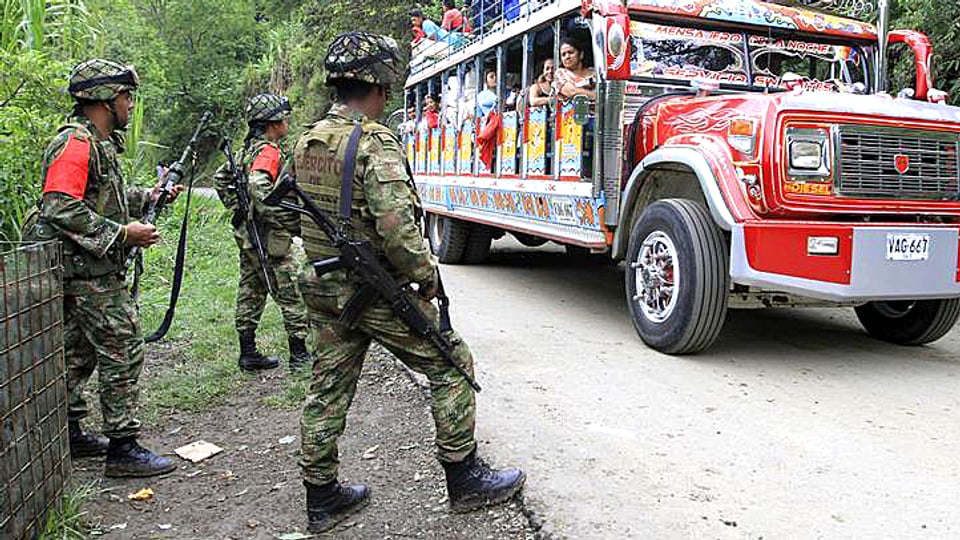 Kolumbianische Regierungssoldaten stehen an einer Dschungelstrasse. Ein farbiger Reisebus passiert die Patrouille.