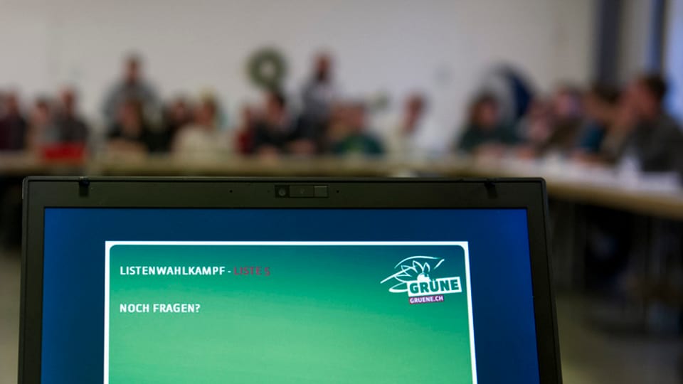 Laptop mit Grünen-Logo, im Hintergrund Parteiversammlung