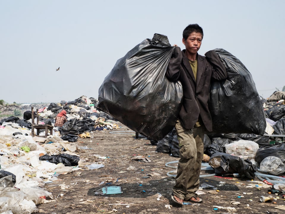 Ein Mann geht über eine Müllhalde, links und rechts trägt er vollgepackte Müllsäcke. 