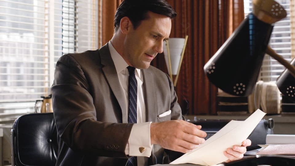 Don Draper (Jon Hamm) an seinem Bürotisch, in der Hand ein paar Papiere.
