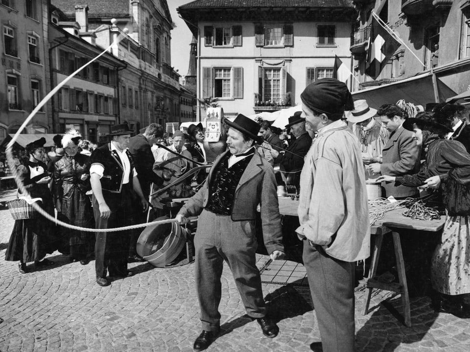 Szene auf einem Marktplatz. Ein Mann knallt eine Geissel. Ein zweiter schaut ihm dabei zu. Im Hintergrund stellen sich Leute an einem Stand an.