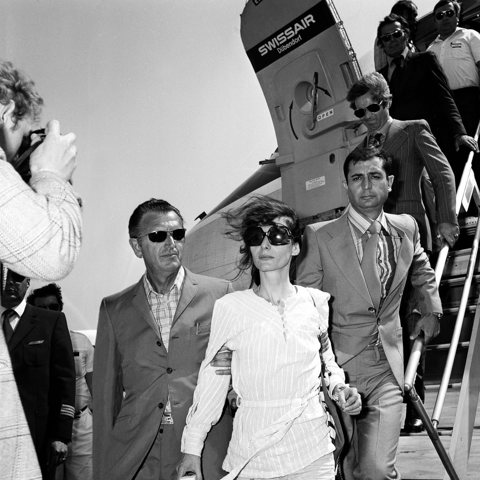 Eine dünne Frau mit Sonnenbrille steigt aus einem Flugzeug und wird links und rechts von Männern festgehalten. 