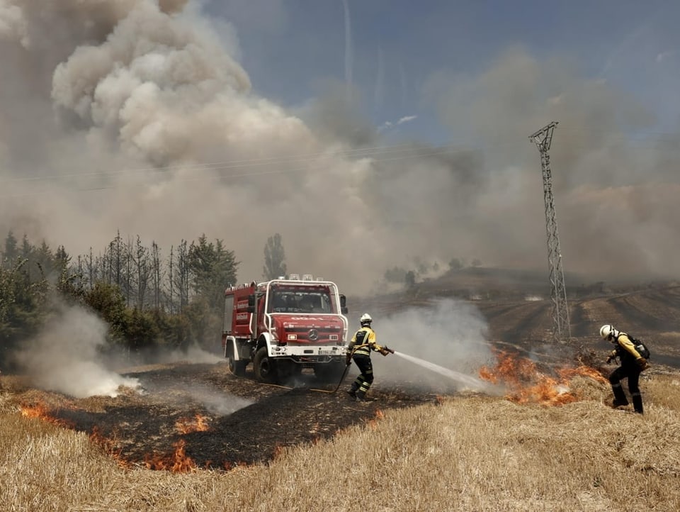 Zwei Feuerwehrleute löschen einen Brand auf einem ausgetrockneten Feld. Im Hintergrund steht ihr Fahrzeug.