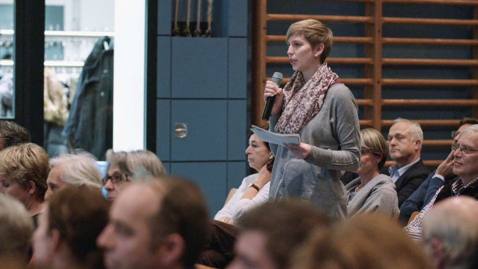 Eine junge Frau steht mit einem Mikrofon bei einer Versammlung zwischen sitzenden Menschen.