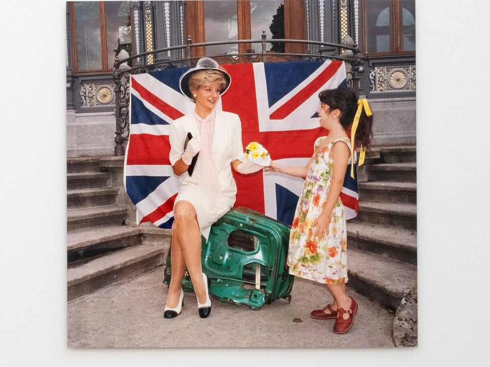 Prinzessin Diana auf einem Autowrack vor einer britischen Flagge, neben ihr ein Mädchen, das ihr Blumen reicht.