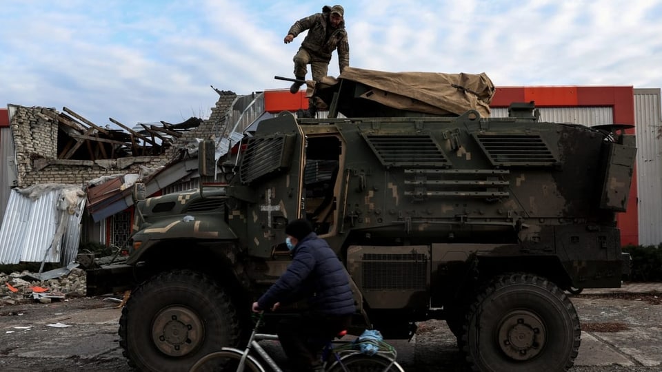 Ein Soldat steht auf einem Militärfahrzeug, davor fährt ein Mann auf einem Velo vorbei.