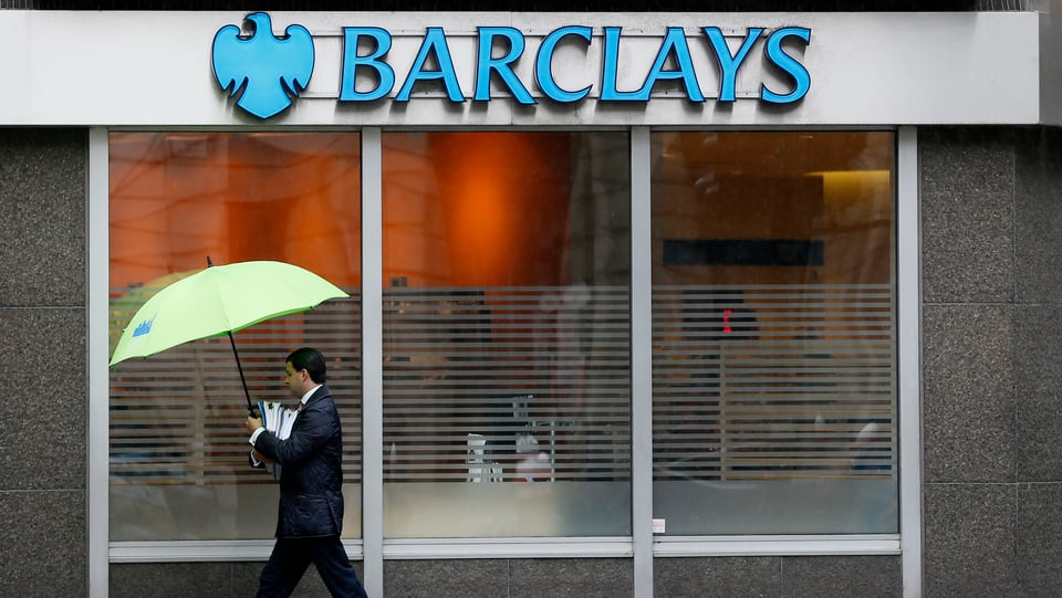 Ein Banker in Anzug und mit Regenschirm läuft an einer Filiale der Barclays Bank vorbei.