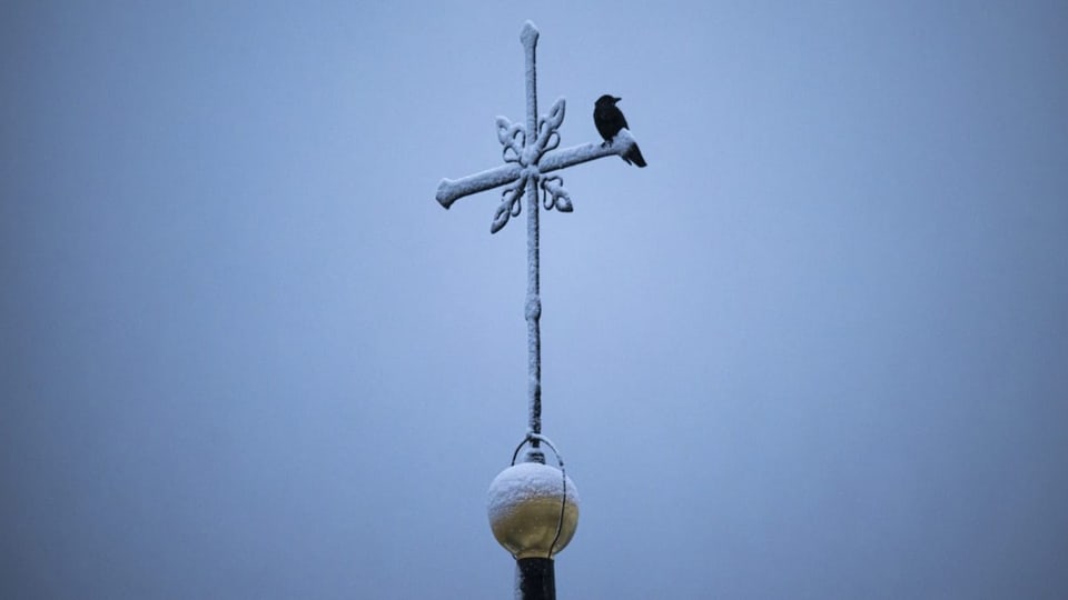 Ein Vogel sitzt auf einem verschneiten Kirchturmkreuz.