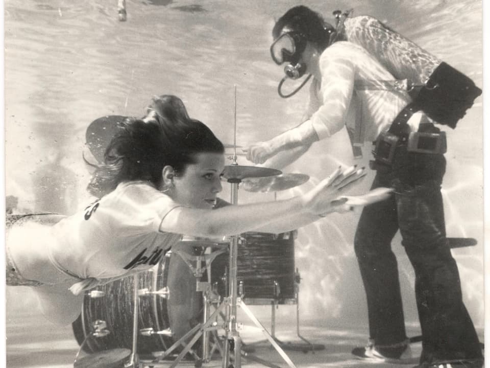 Schwarz-weiss-Bild eines Schlagzeugers, der mit Atemmaske unter Wasser spielt, im Vordergrund schwimmt eine Frau im T-Shirt vorbei. 