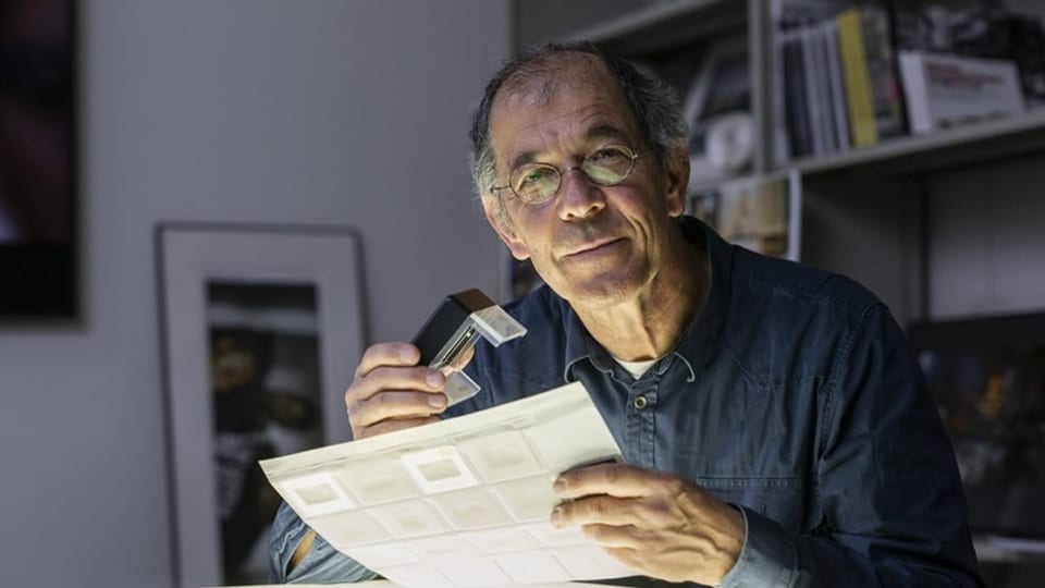 Mann mit grauen Haaren, Brille und blauem Hemd sitzt in einem Atelier und schaut sich Dias an