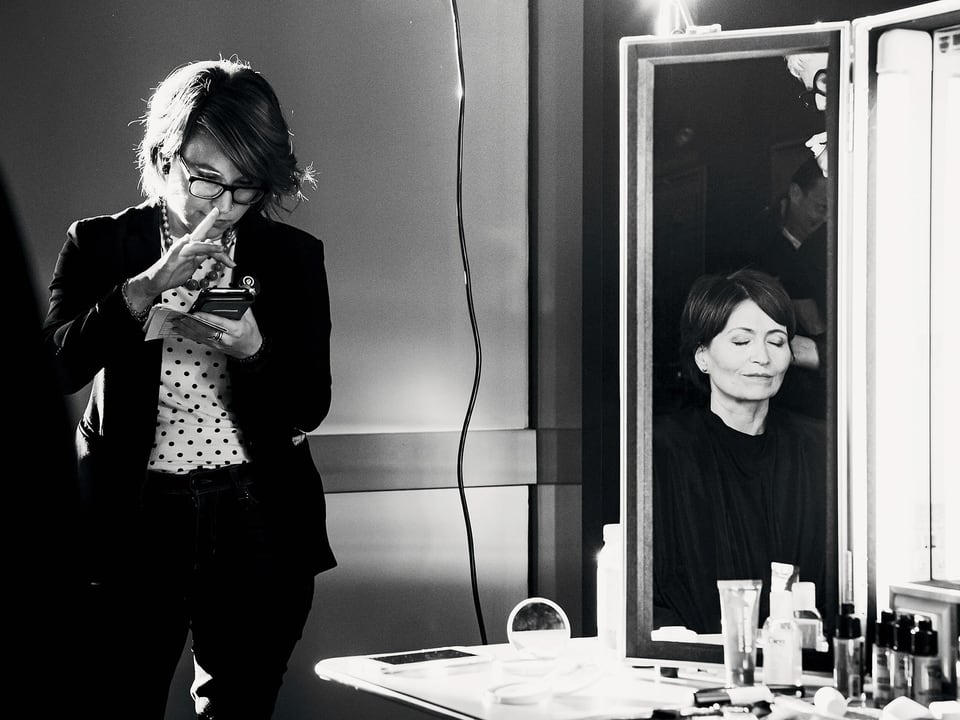 Schwarzweissbild: Eine Frau vor beleuchtetem Schminkspiegel, neben ihr stehend eine Frau am Smartphone