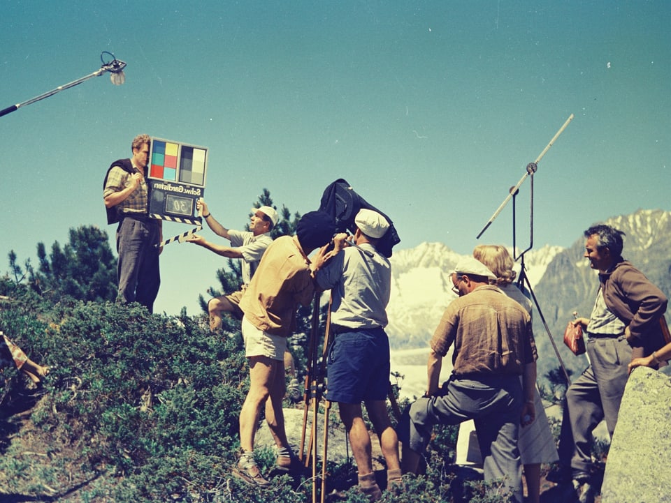 Hannes Schmidhauser steht auf einer Anhöhe auf der Alp in Mitten von Heidelbeersträuchern. Vor ihm wird die Klappe geschlagen. Über ihm hängt die Tonangel, vor ihm stehen mehrere Crewmitglieder und die Kamera sowie ein Lichtreflektor.