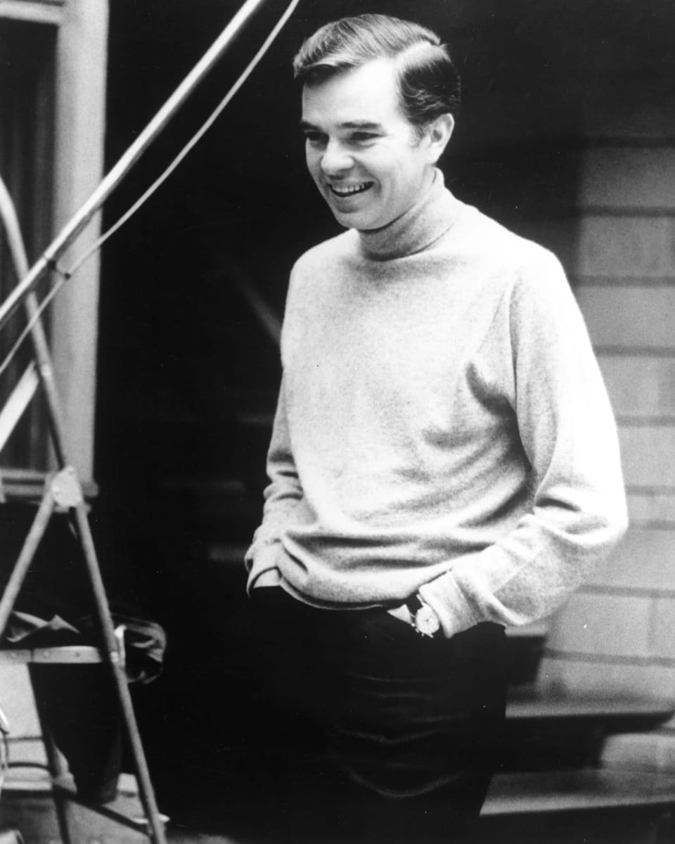 Schwarzweiss-Foto eines jungen Mannes in hellem Rollkragenpulli mit Seitenscheitel, die Hände in den Hosentaschen
