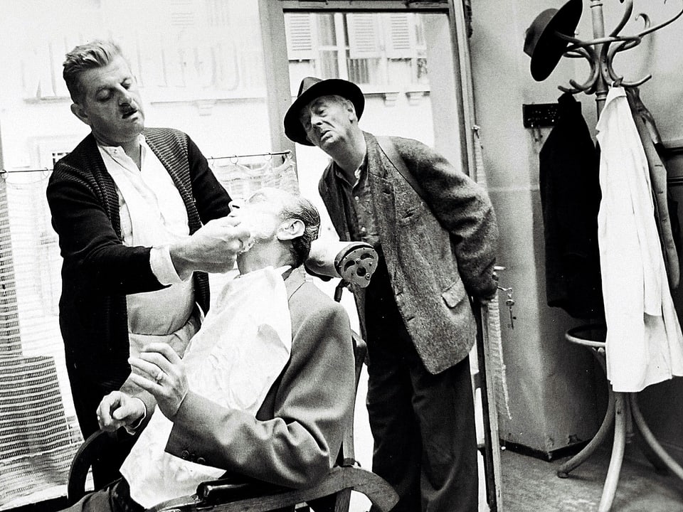 Ein Mann steht vor dem Friseurstuhl und rasiert einen Kunden. Dahinter steht ein dritter Mann und sieht zu.