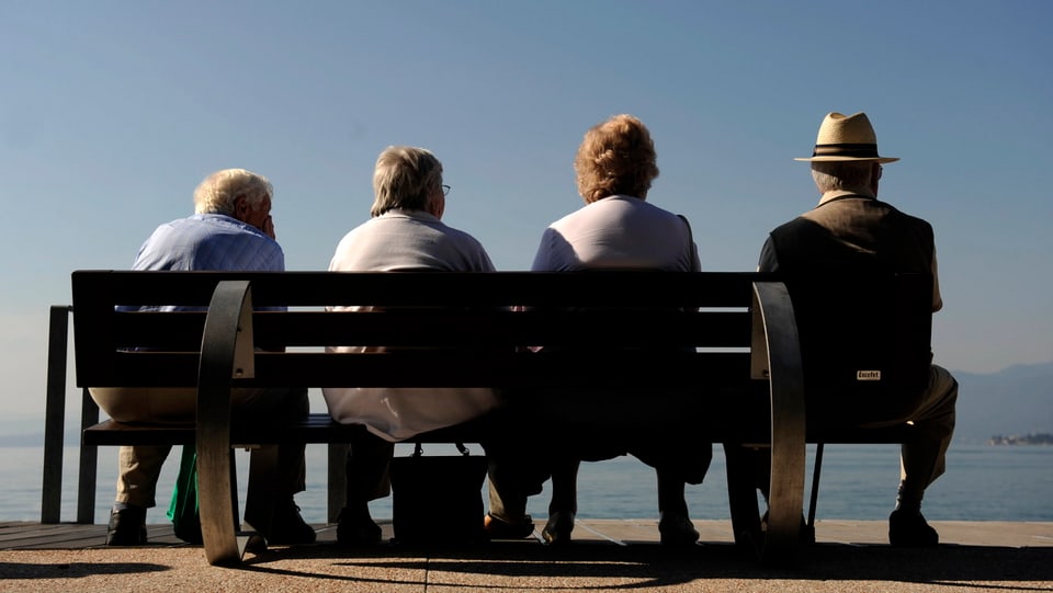 Vier ältere Personen sitzen auf einer Bank mit dem Rücken zur Kamera und blicken auf einen See.