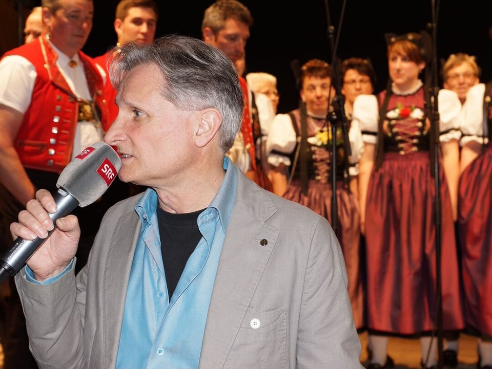 Moderator mit MIkrofon vor Jodelchor im Hintergrund.