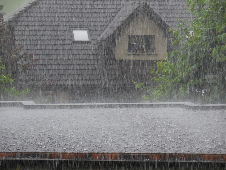 Intensiver Starkregen prassel auf ein Garagendach, im Hintergrund ein steiles Hausdach.