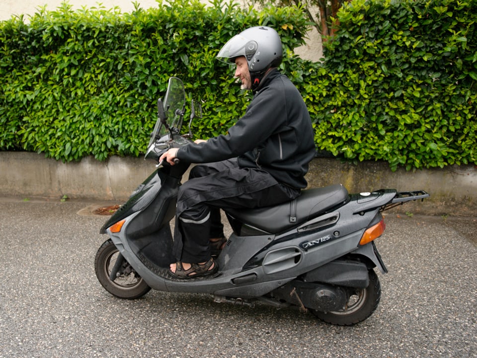 Roman Leonardi mit Helm, dunklem Regenschutz und Sandalen auf seinem Roller.