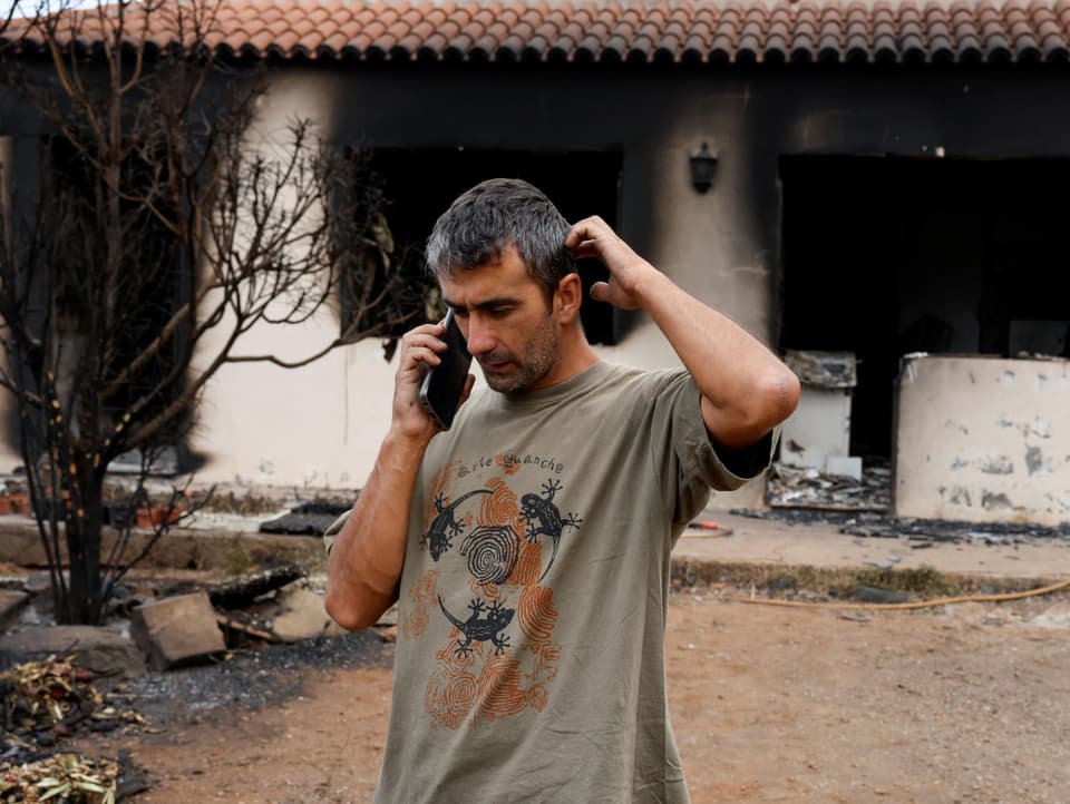 A man standing with a phone in front of a burning house.  The wall next to the windows is black.