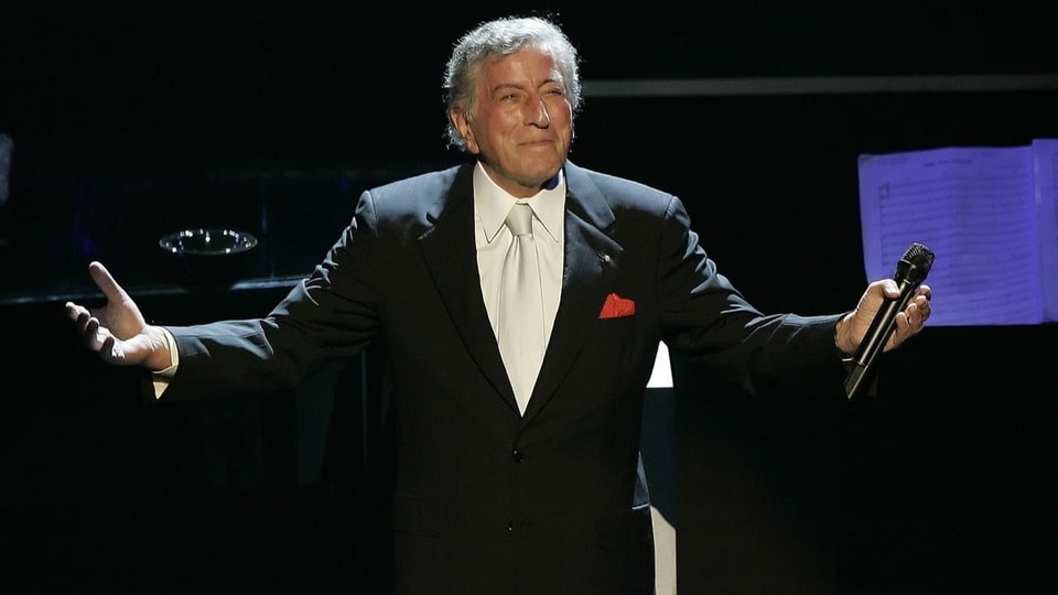 Tony Bennett stands on stage holding a microphone and arms outstretched.