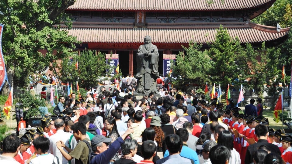 Menschen stehen dicht an dicht vor einem Tempel. In der Mitte steht eine Konfuzius-Statur