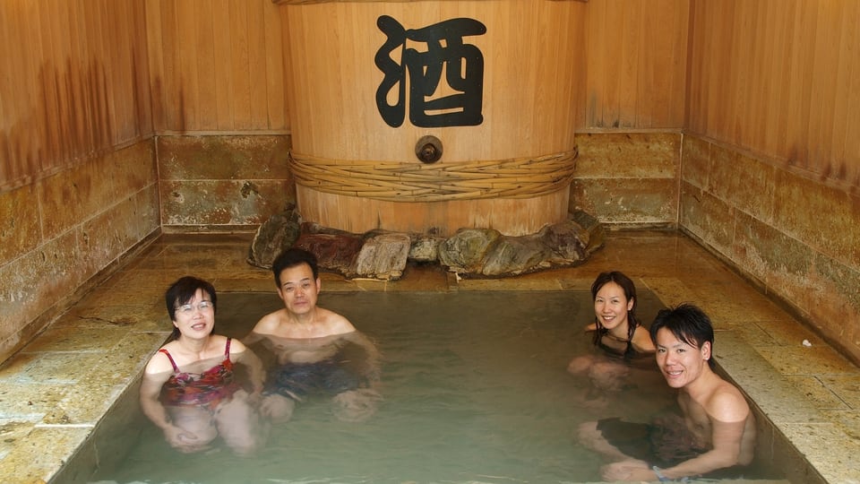 Eine Gruppe von vier jungen Japanerinnen und Japanern sitzen in einem kleinen, quadratischen Sake-Bad vor einem grossen Holzfass mit dem japanischen Reiswein.