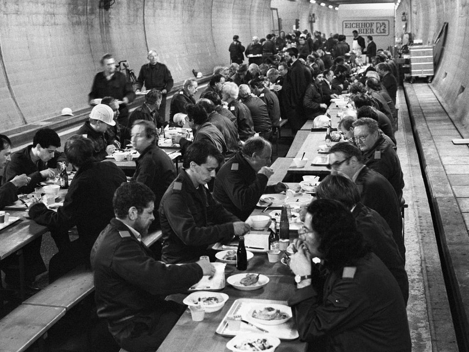 Zu sehen sind etwa 50 Menschen, die in einem Tunnel an langen Tischen und Bänken sitzen und essen. 