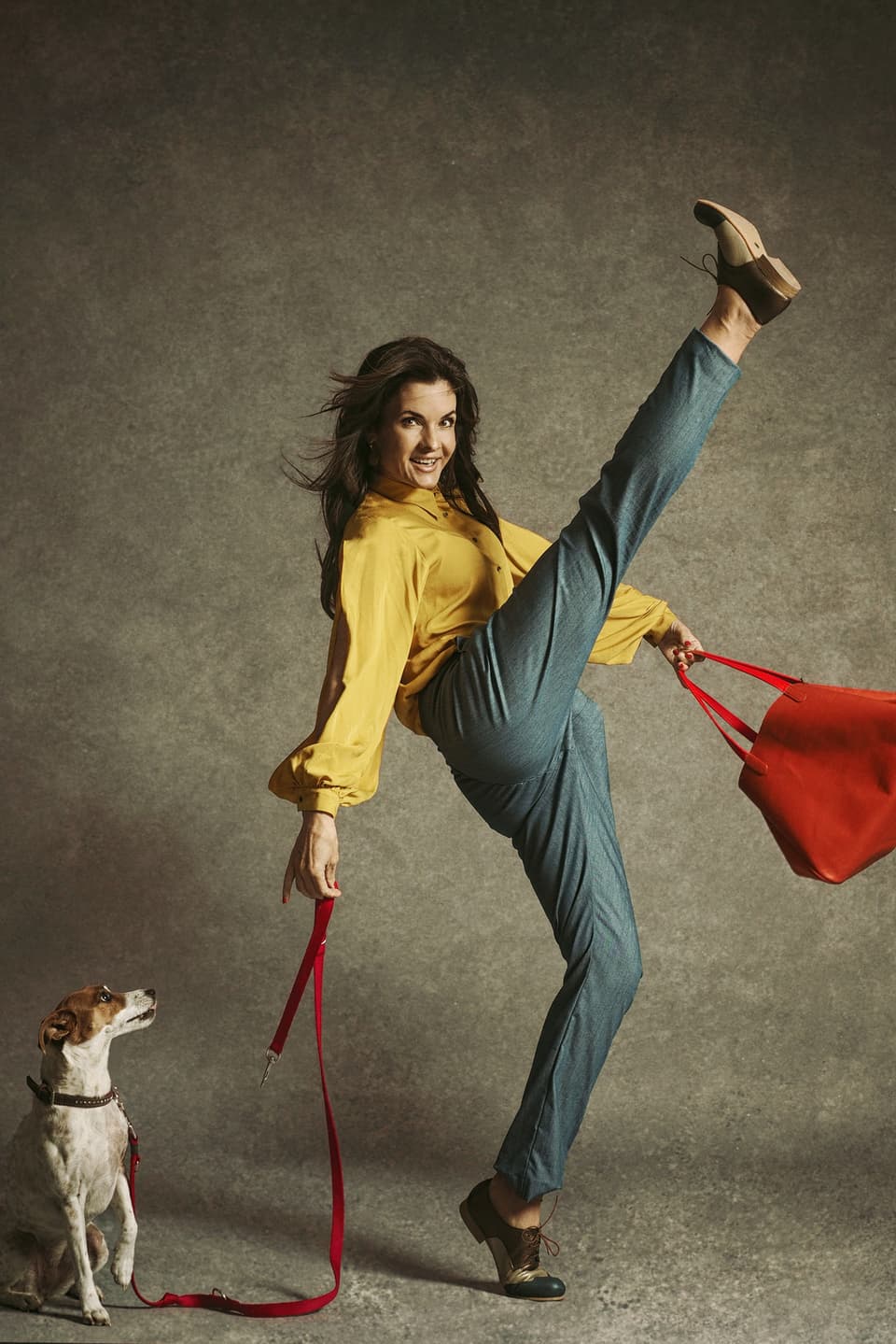 Eine Frau hebt ihr Bein durchgestreckt in die Höhe. In der einen Hand trägt sie eine Tasche, in der anderen hält sie einen Hund an der Leine.