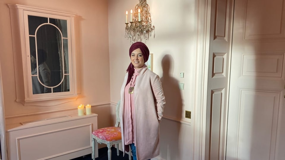 Fatima Zibouh steht in einem Zimmer und trägt rosarote Kleider. Dazu ein bordorotes Kopftuch. 