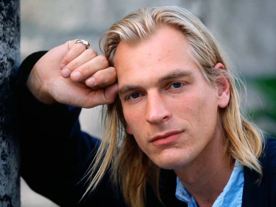 Julian Sands the Younger leans on a pole and faces the camera.