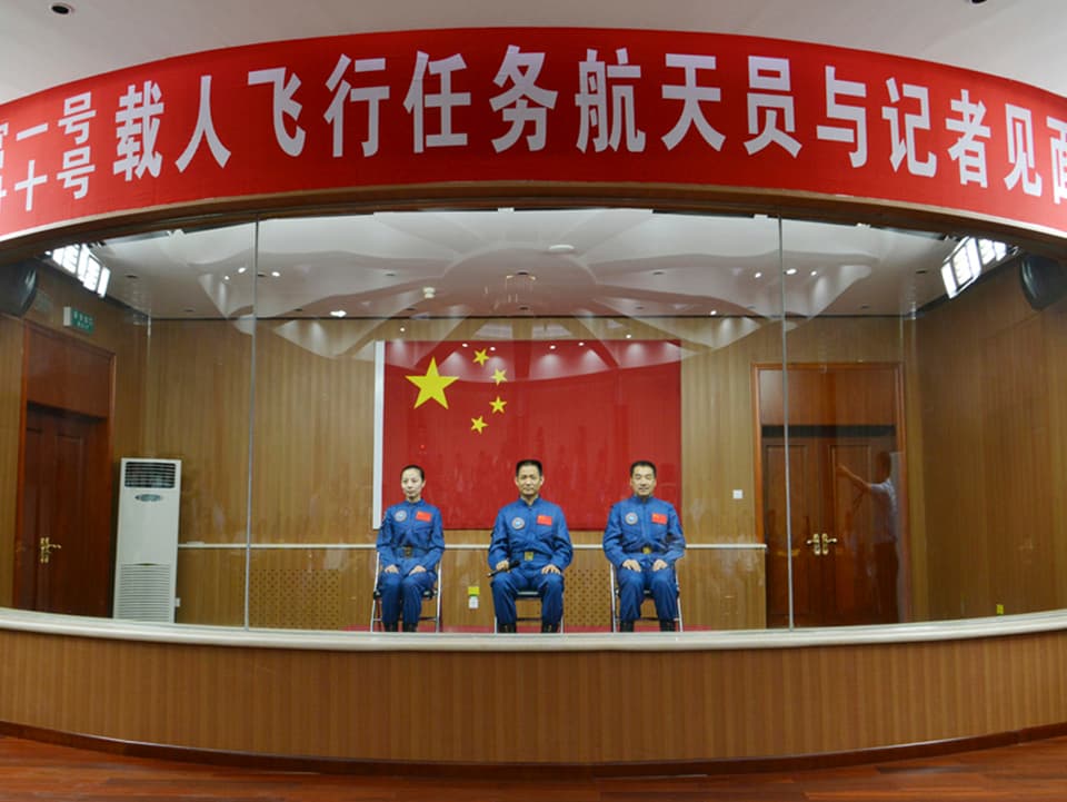 Drei Astronauten sitzen nebeneinander auf Stühlen in einem Glaskasten mit chinesischen Schriftzeichen.