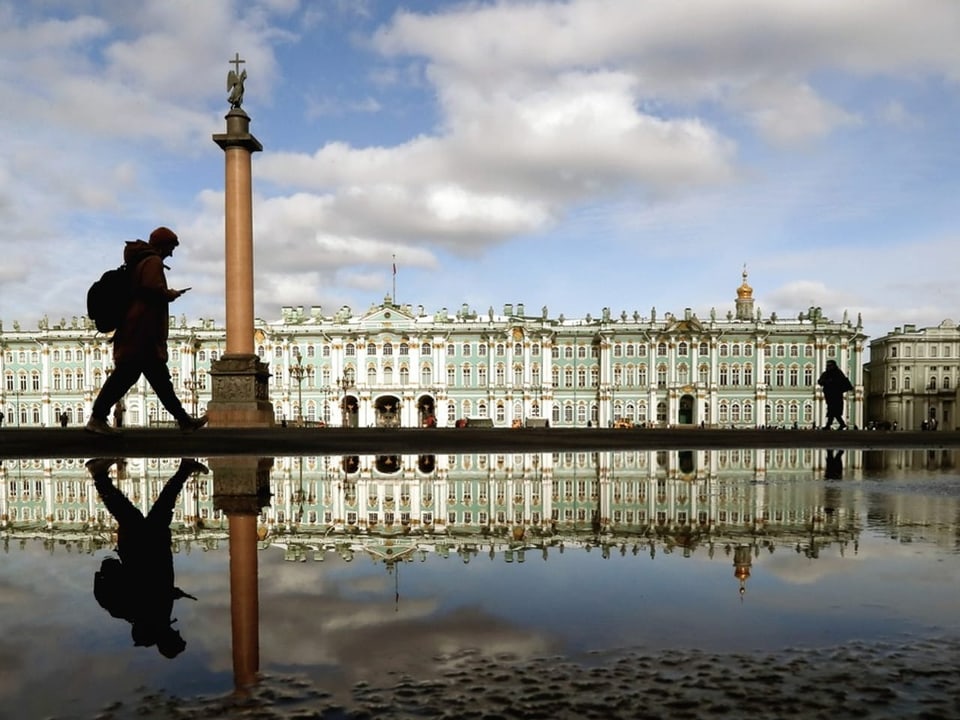 Ein Mann überquert einen Platz in St. Petersburg. Er, eine Säule und ein Gebäude spiegeln sich in einer Pfütze