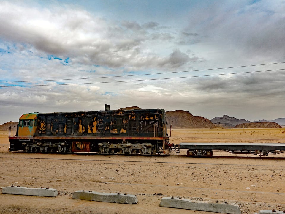 Eine alte Güter-Lokomotive mit abblätternder Farbe steht in einer Wüste. Im Hintergrund sieht man weit entfernt Felsen.
