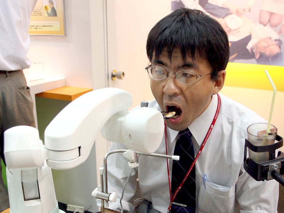 Ein Japaner sitzt mit offenem Mund vor einer weissen Maschine, die ihm einen Happen Essen in den Mund schiebt.