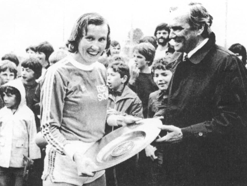 Eine Fussballerin erhält von einem Mann einen Pokal, im Hintergrund freuen sich Kinder.
