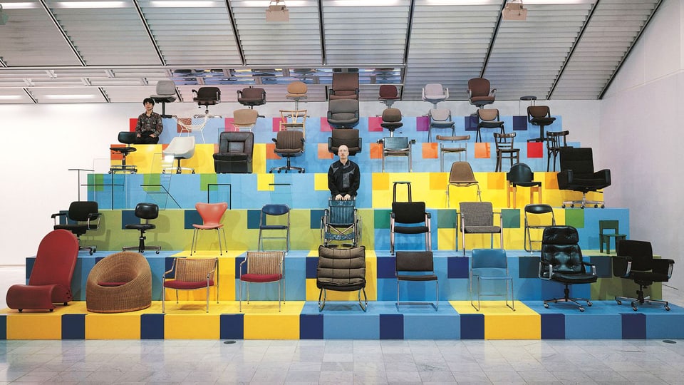 Auf einem Podest in verschiedenen Farben sind Stühle aller Art ausgestellt, dazwischen steht Christian Philipp Müller selber.