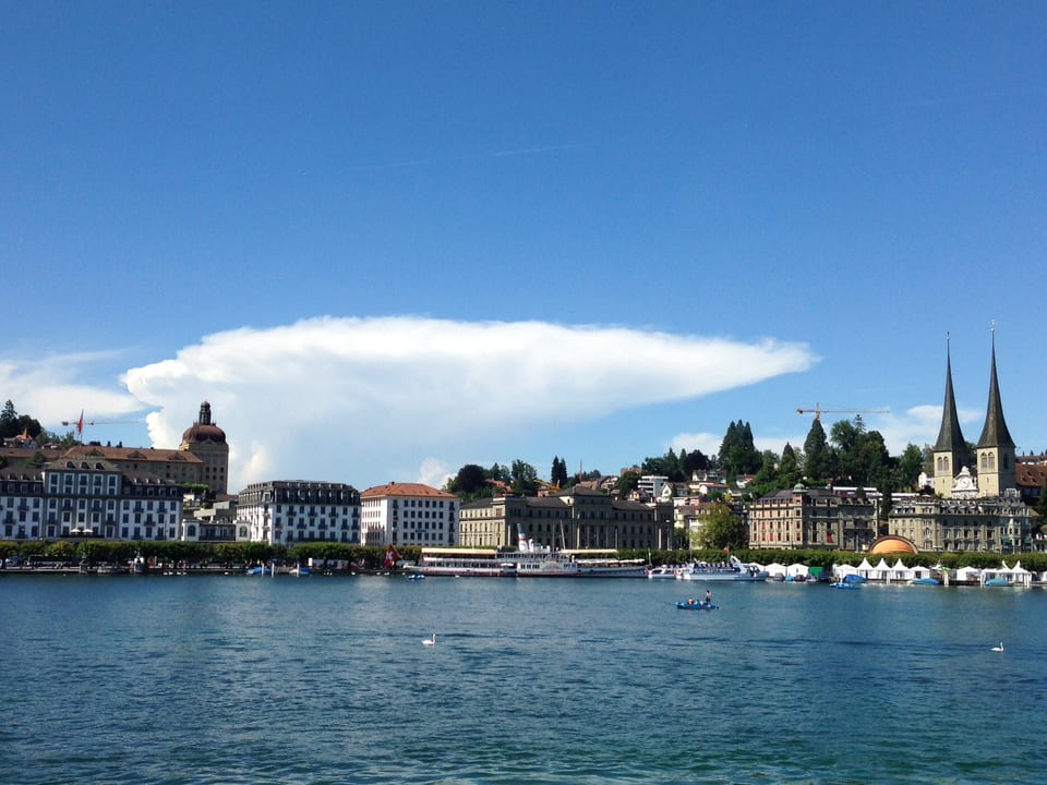 See, dahinter Ufer von Luzern mit Häusern und Kirche. Der Himmel ist blau, aber ein einsamer Gewitteramboss ist am Horizont zu sehen. 