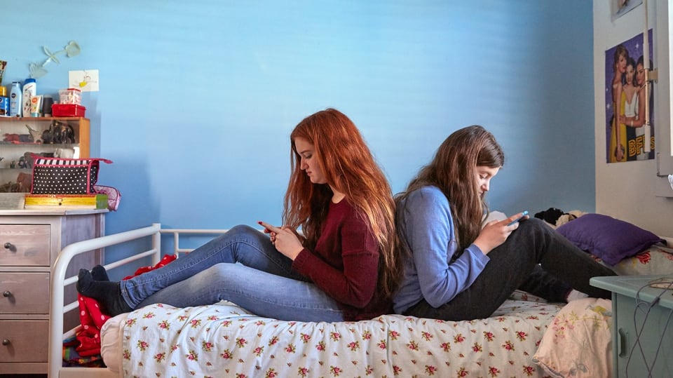 Zwei Mädchen hängen am Smartphone, Rücken an Rücken auf einem Bett im Kinderzimmer.
