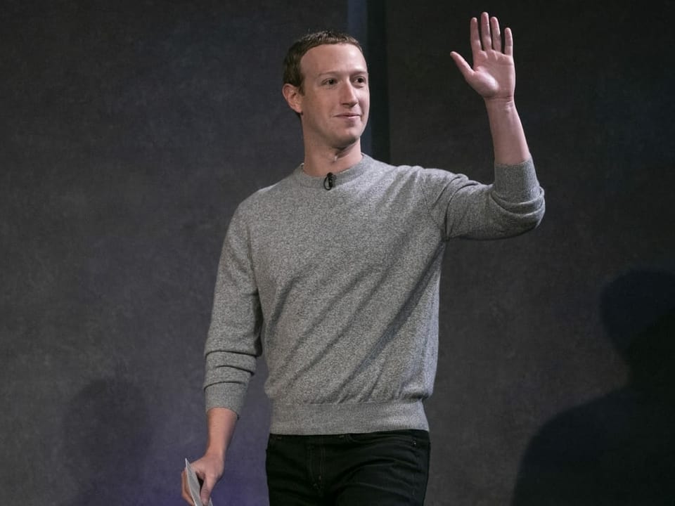 Zuckerberg lächelt und winkt mit der linken Hand.