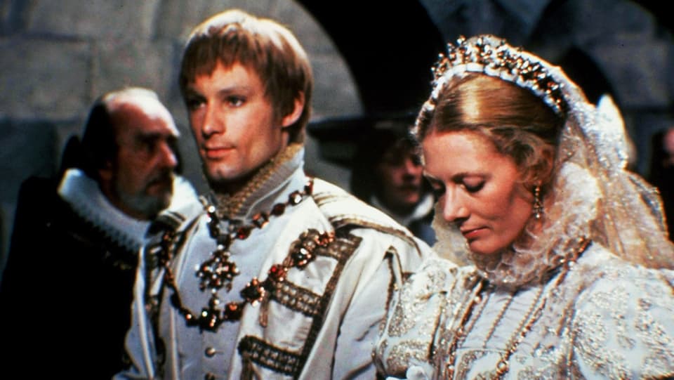 Eine Szene aus einem Film: Eine Frau und ein Mann stehen nebeneinander und haben die Hände zusammengelegt wie zum Gebet. Sie sind prunkvoll gekleidet, mit Krone, Ketten etc.