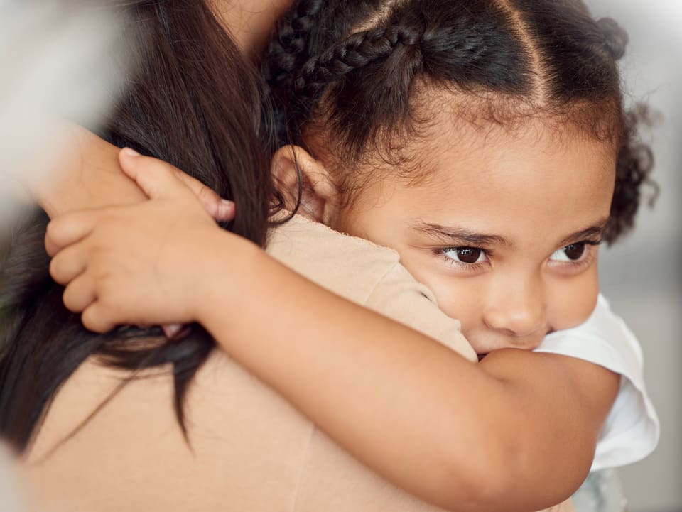 Ein junge Mädchen mit geflochtenen Zöpfen umarmt ihr Mutter, die man nur von hinten sieht. 