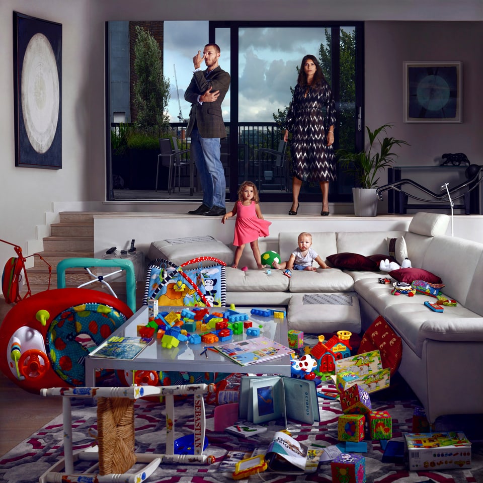 In einem Wohnzimmer türmen sich Kinderspielzeuge. Auf dem Sofa zwei kleine Kinder. Auf einer Ebene darüber stehen ein Mann und eine Frau. 